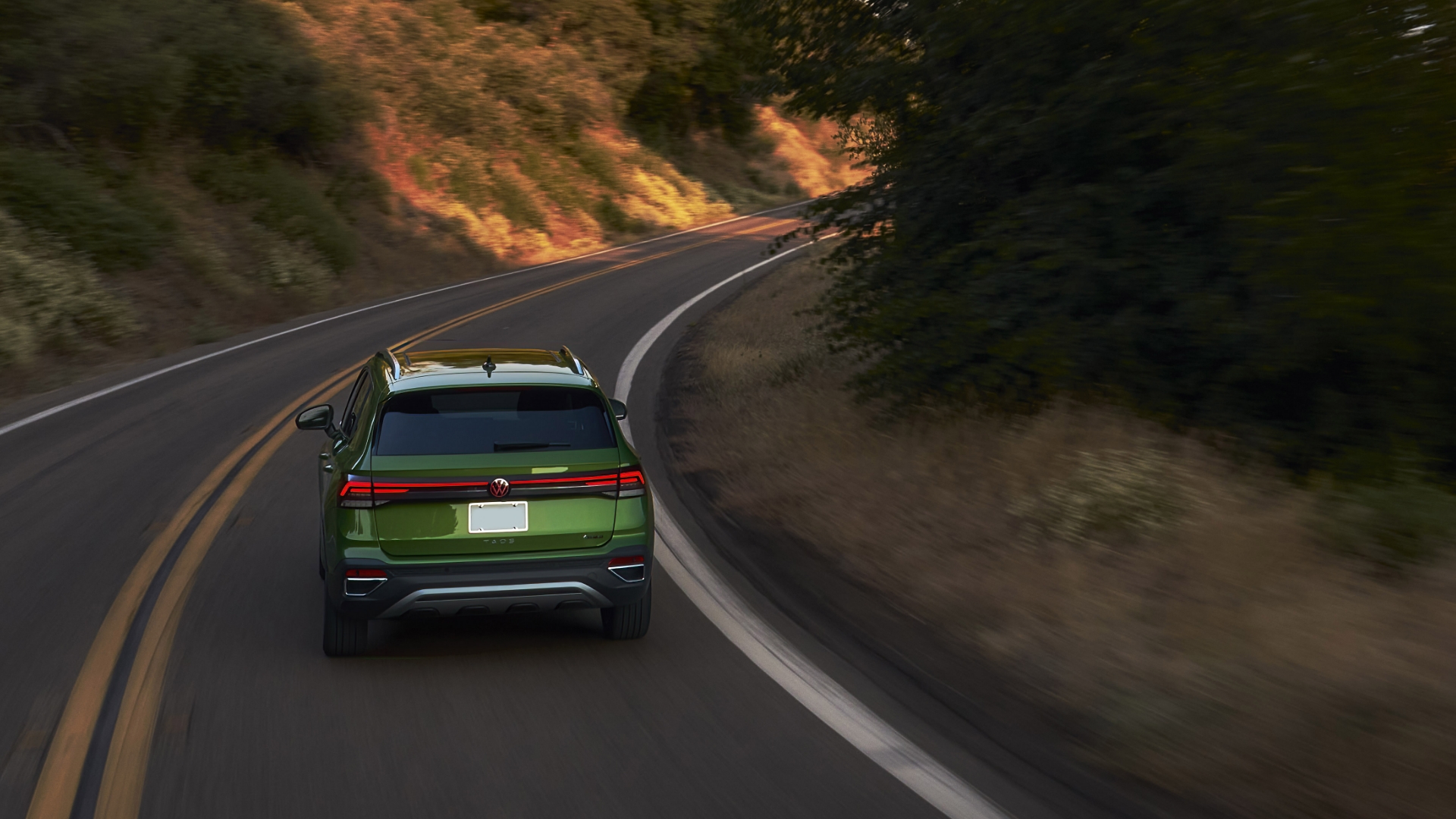 Rear view of a green 2025 Volkswagen Taos driving along a scenic road at sunset.