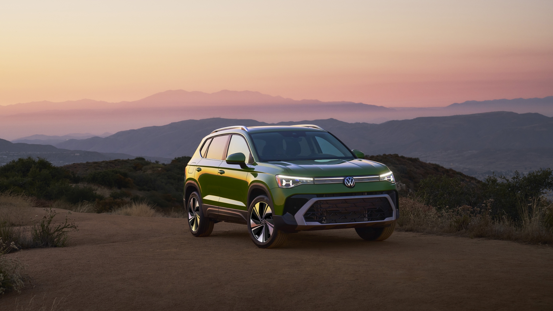 A green 2025 Taos parked on a dirt road during sunset against a backdrop of rolling hills and distant mountain ranges.