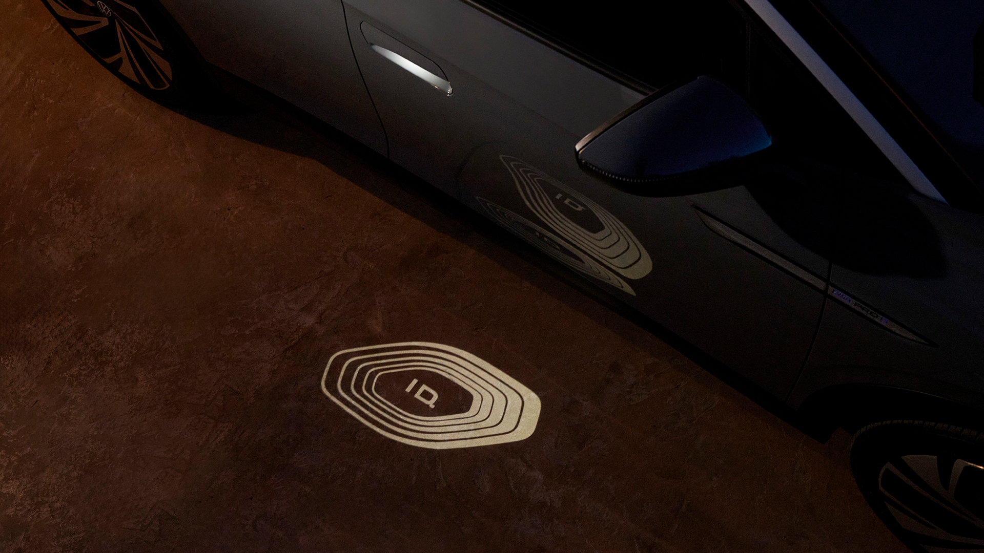 A Volkswagen ID. logo is projected onto the ground from the door of a Volkswagen ID. model vehicle at night.