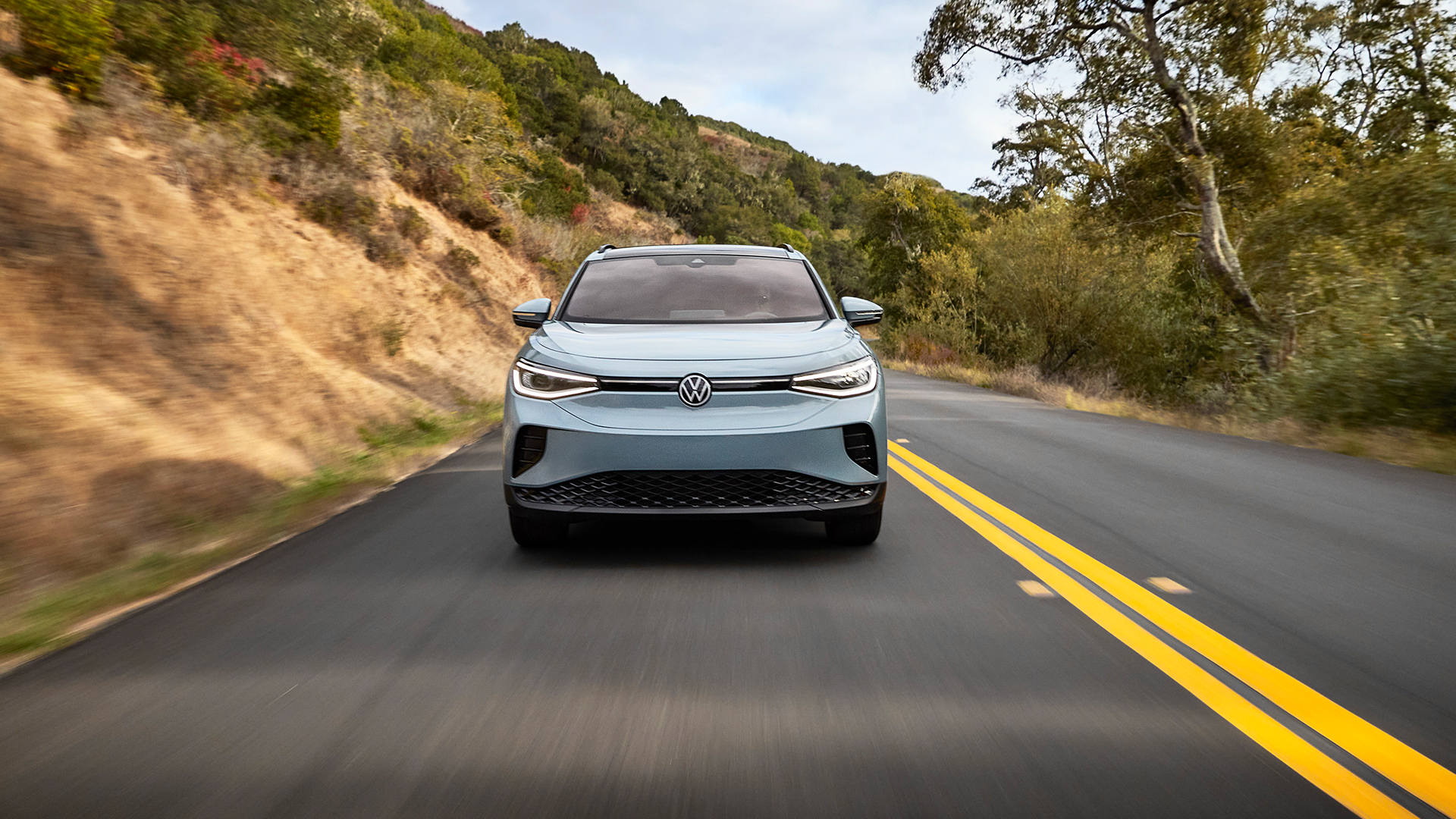 Front view of a silver Volkswagen ID.4 driving on a scenic road through hills.