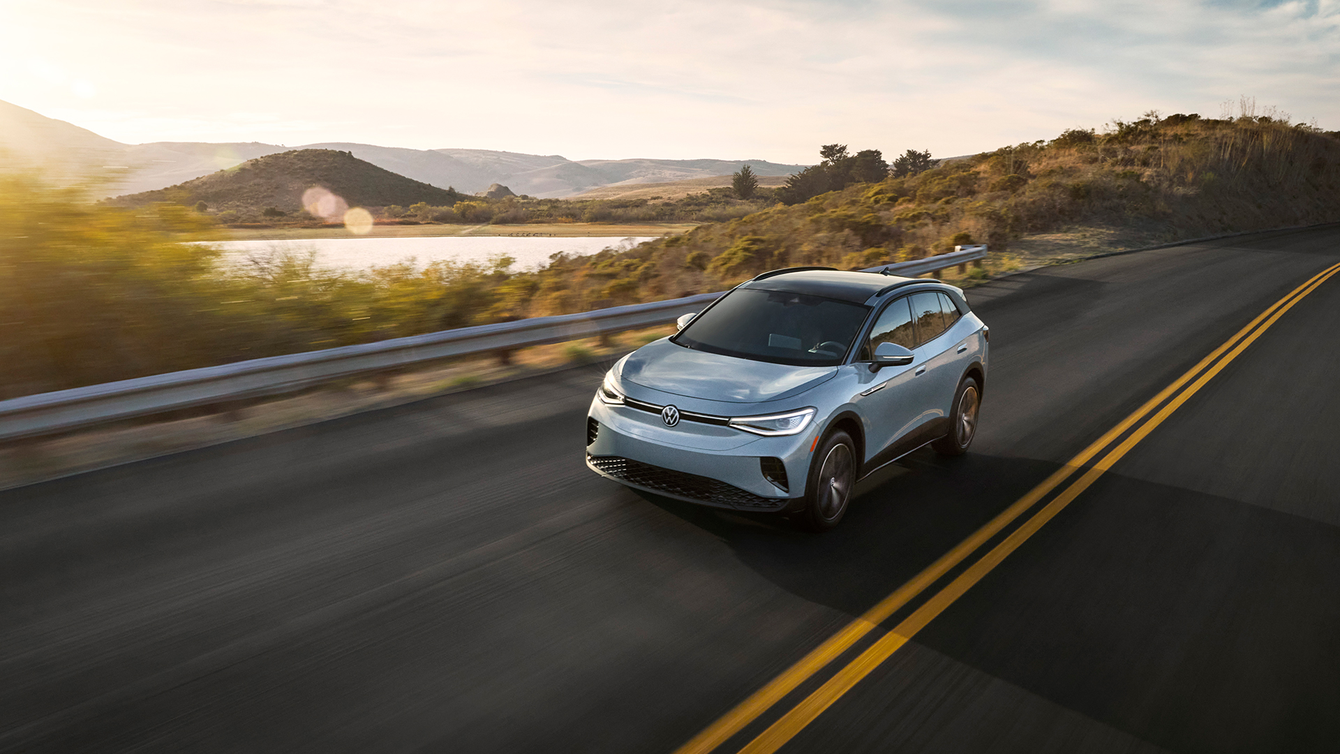 A grey Volkswagen ID.4 Pro electric SUV driving along a highway with a lake and hills in the background.