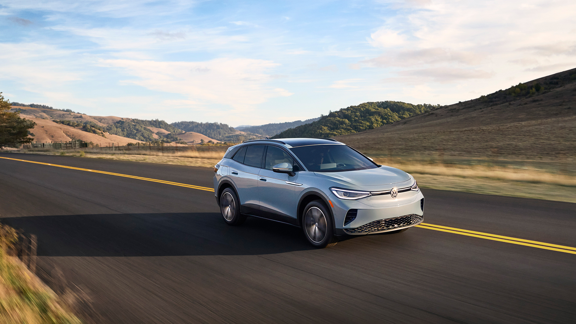 A light gray Volkswagen ID.4 electric SUV, driving on a scenic road with rolling hills in the background.