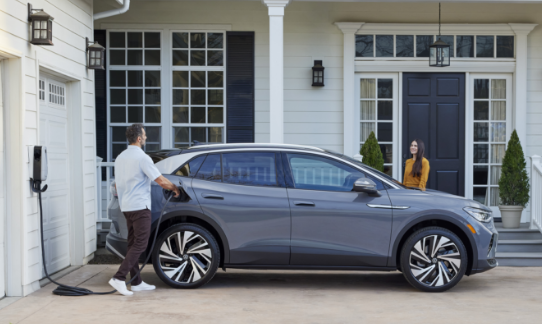 A man is plugging the pure gray Volkswagen ID.4 into a home charging station, while a woman stands beside the car parked by the white house